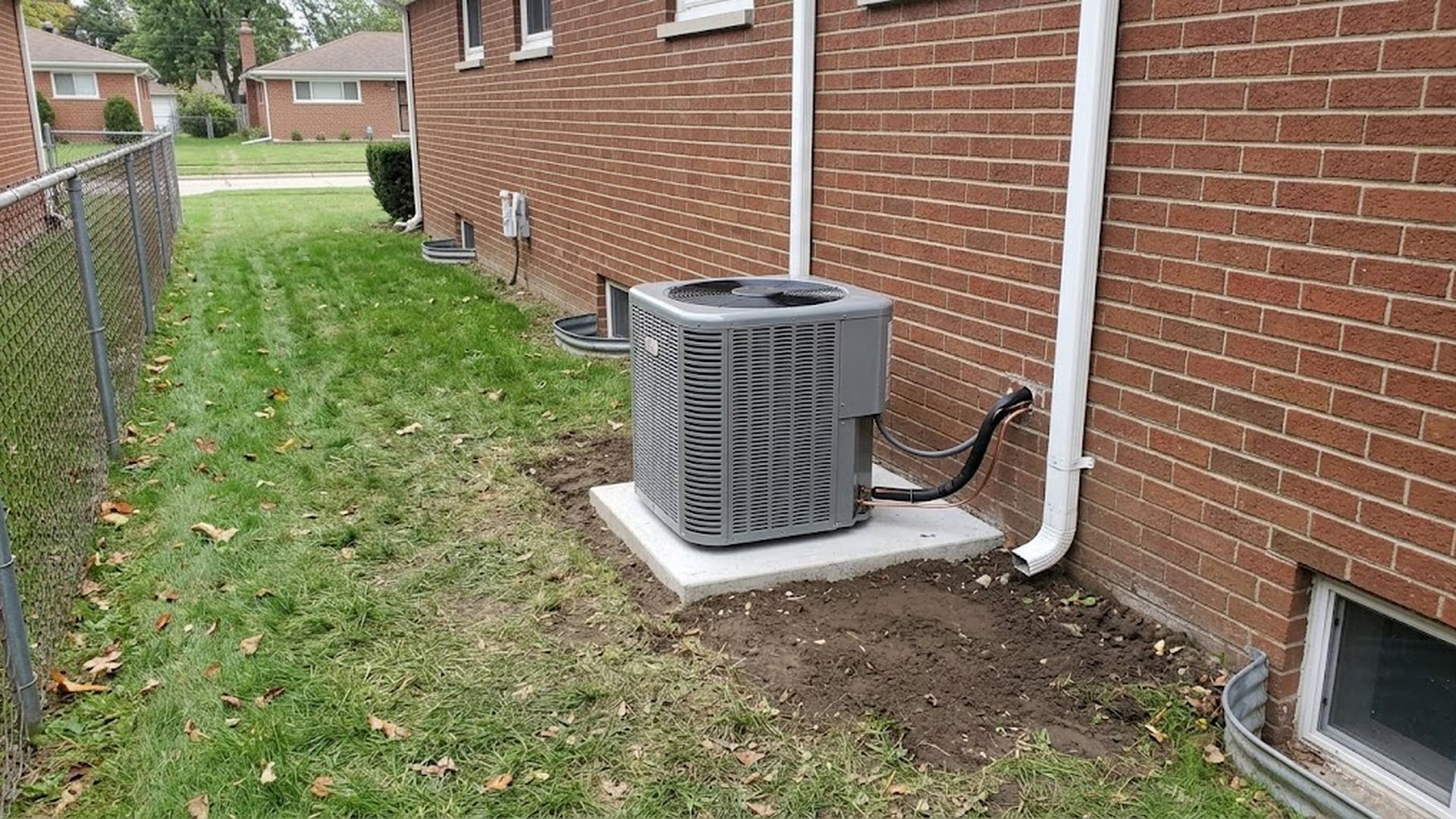 Gray outdoor air conditioner on a concrete pad beside a brick house wall, with a fenced grassy side yard and neighboring houses behind it