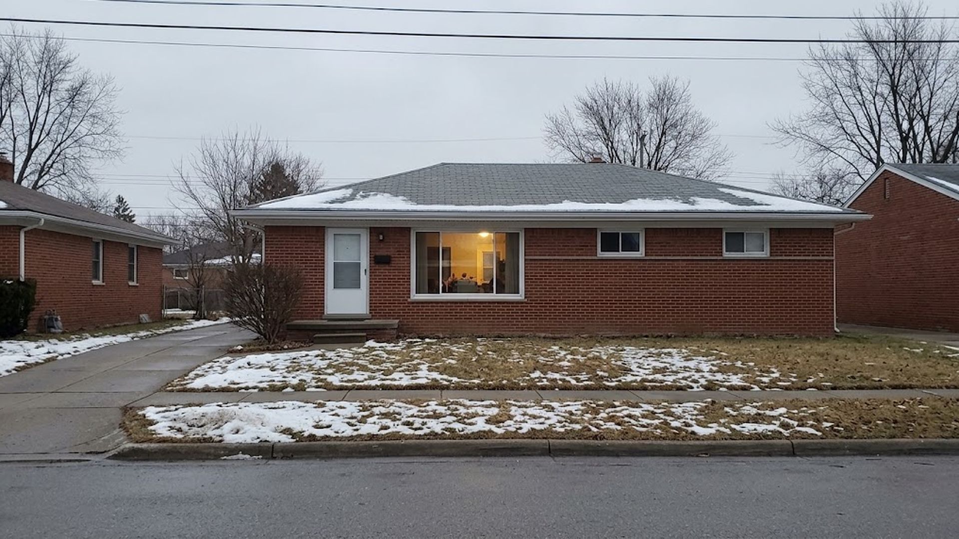Brick ranch house on a residential street on an overcast winter day, with light snow on the lawns and a lit front window