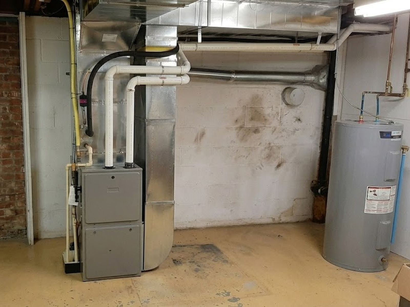 Furnace with white pipes and metal ductwork in a basement utility room, with a water heater against the wall beside it
